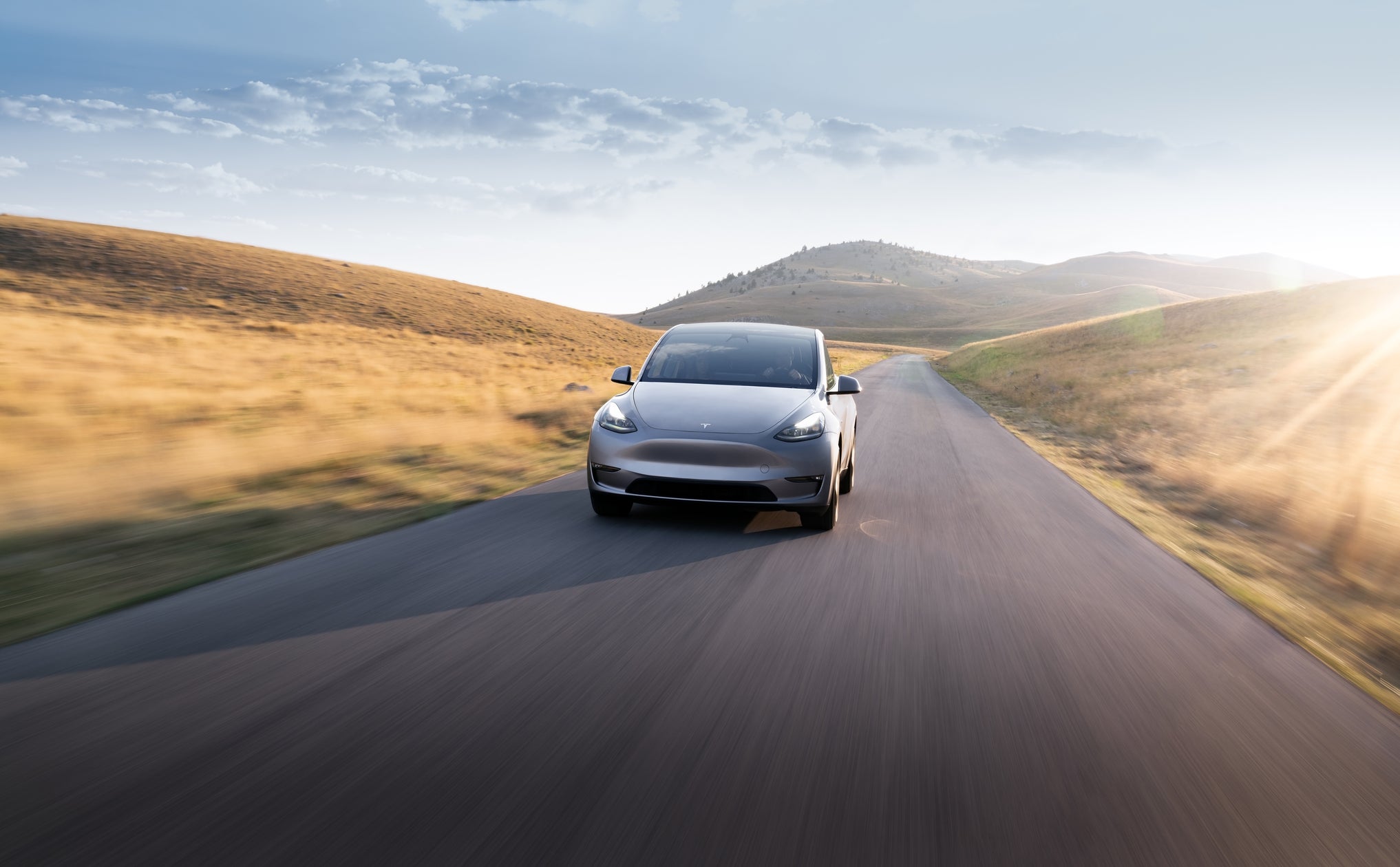 A silver Tesla Model Y driving through sunlit rolling hills